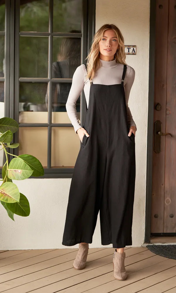Woman in a black linen bamboo jumpsuit over a cream turtleneck, hands in pockets, on a porch beside a window